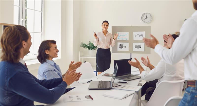 Team of professionals in a meeting room applauding a presenter, who is standing and explaining data displayed on charts behind her.